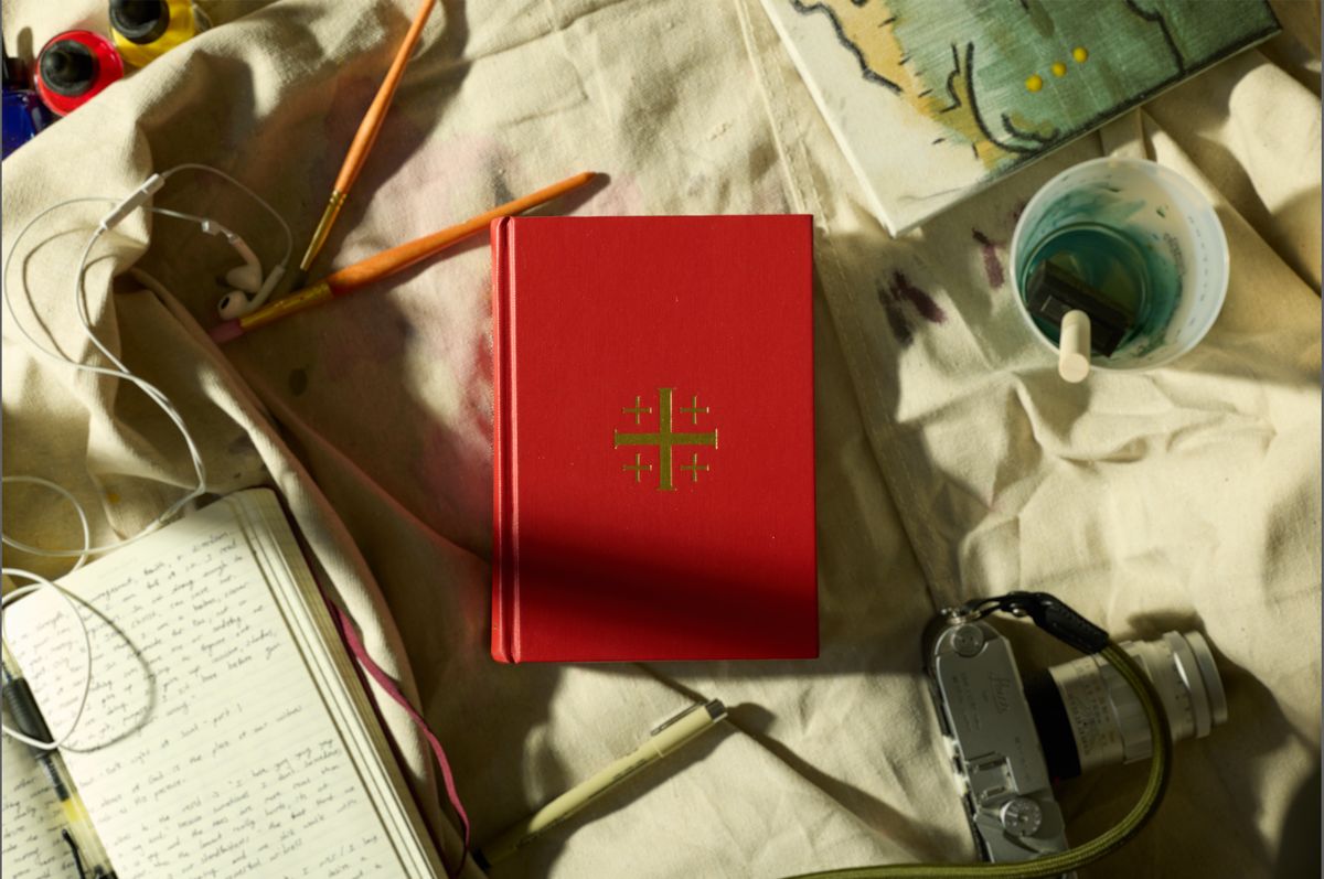 A red prayer book with a gold Jerusalem cross on a paint-stained canvas cloth, surrounded by brushes, a handwritten journal, a film camera, and a cup of rinse water